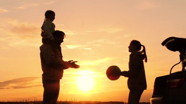 Family playing with ball in nature at sunset, silhouettes. Daughter sits on father's shoulders, throwing ball with mother. Family travel adventures by car, active leisure. Weekend road trip to nature.