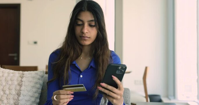 Serious attractive young Indian woman using bank application on smartphone, holding credit card, typing, making money transaction, paying for purchase on webstore, buying goods on Internet shop