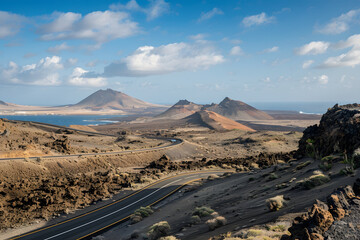 A scenic road meanders through a stark volcanic landscape under a blue sky with fluffy clouds, evoking adventure and exploration.