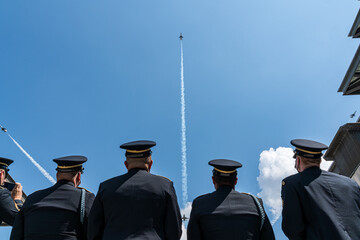 A group of men in military uniforms are watching a jet fly in the sky - obrazy, fototapety, plakaty