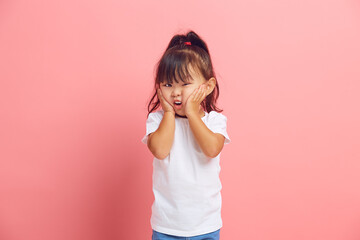 Puzzled little asian girl feels fear, holds her hands on cheeks, looks with great surprise and amazement at the camera standing on a pink isolated background