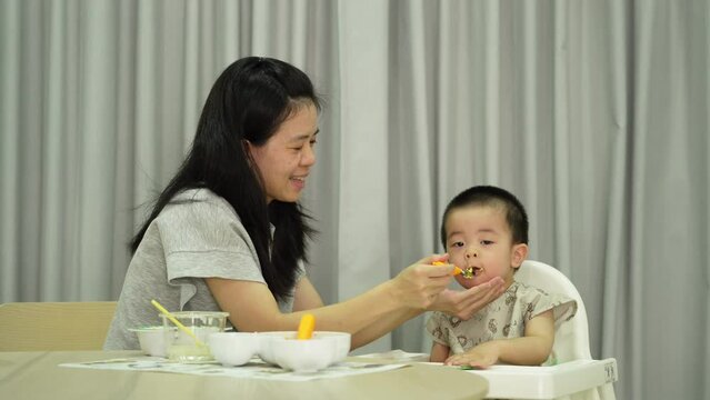Asian mother feeding baby boy sitting on white highchair with messy table and gray backdrop