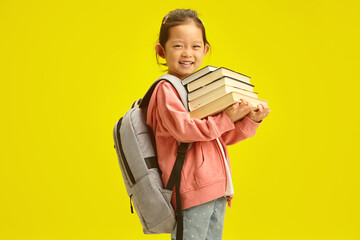 Cheerful little korean first grader girl in casual clothes with many books for studying at school and backpack on shoulders isolated over yellow background - 785532494