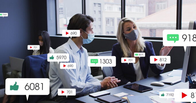 Image of social media icons over diverse man and woman wearing face masks discussing at office