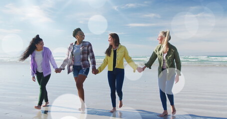 Spots of light against group of diverse female friends holding hands walking on the beach