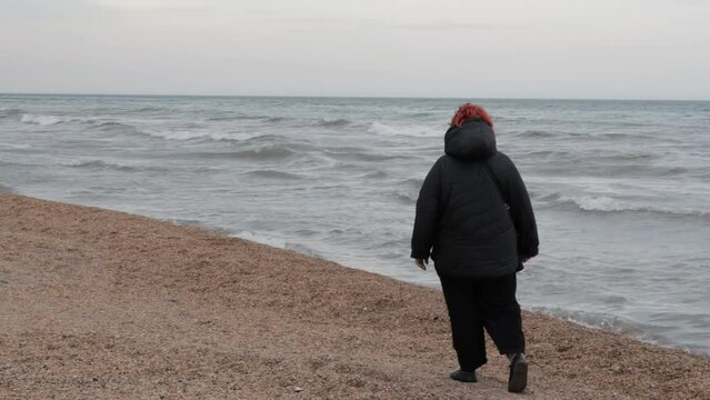 A walk in cool windy weather by the sea. An adult woman walks along the beach in warm clothes. A beach with small shells on the shore of the Caspian Sea. Waves at sea and strong wind.