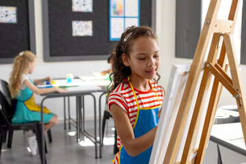 In school, during art class, a young biracial girl wearing a striped apron is painting
