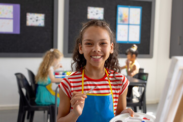 In school art class, biracial young girl holding a paintbrush smiles at camera