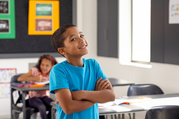In school, young biracial boy standing with arms crossed, looking thoughtful in the classroom - Powered by Adobe