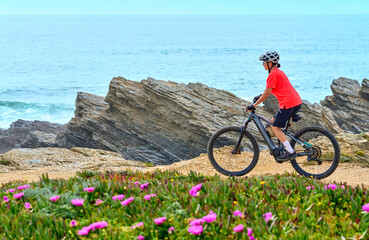 nice senior woman riding her electric mountain bike at the rocky and sandy coastline of the atlantic ocean in Porto Covo, Portugal, Europe - 785549086