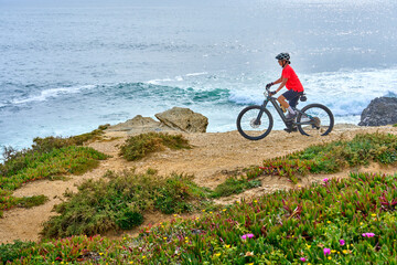 nice senior woman riding her electric mountain bike at the rocky and sandy coastline of the atlantic ocean in Porto Covo, Portugal, Europe - 785549093