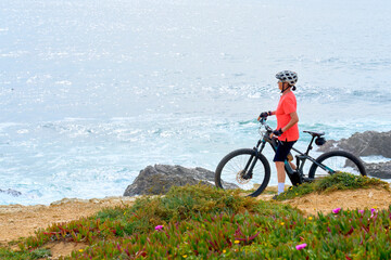 nice senior woman riding her electric mountain bike at the rocky and sandy coastline of the atlantic ocean in Porto Covo, Portugal, Europe - 785549223