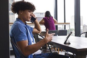 Biracial young man talking on phone, pointing at tablet in a modern business office - 785550004