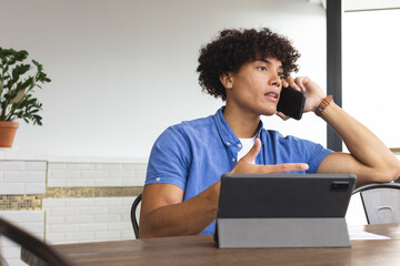 Young biracial man talking on phone, looking away, sitting at table in a modern business office - 785550013