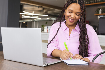 A young biracial woman wearing a pink shirt is writing in a notebook in a modern business office