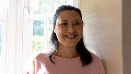 Mature biracial woman standing by window, smiling, at home