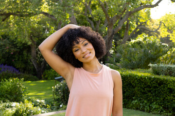 Young biracial woman standing in sunny garden at home, stretching neck, smiling