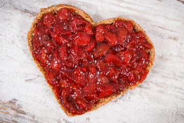 Slice of bread in shape of heart with strawberry jam for breakfast. Rustic background