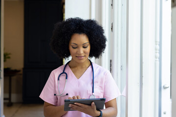 Young biracial female nurse wearing pink scrubs checking tablet at home - Powered by Adobe