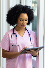 Biracial female nurse wearing pink scrubs reviewing notes on tablet at home