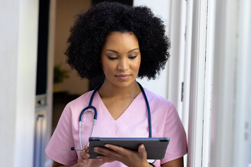 Young biracial nurse wearing pink scrubs checking tablet at home
