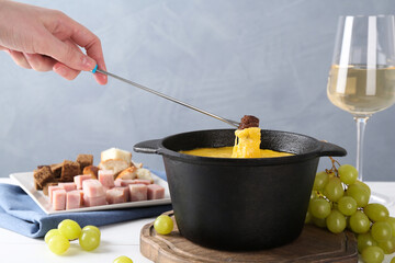 Woman dipping piece of bread into fondue pot with tasty melted cheese at white table against gray...