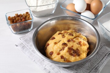 Dough with raisins in bowl and ingredients on white wooden table