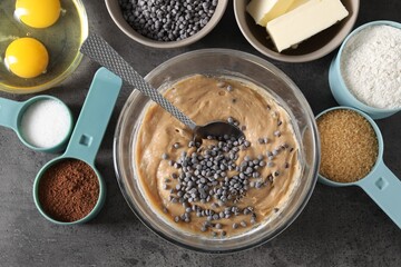 Raw dough with chocolate chips in bowl and ingredients on grey table, flat lay