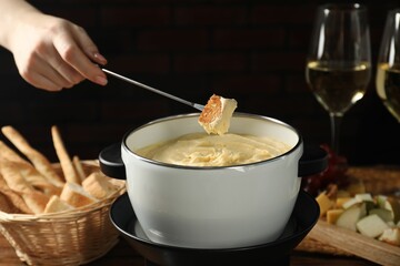 Woman dipping piece of bread into fondue pot with melted cheese at table, closeup