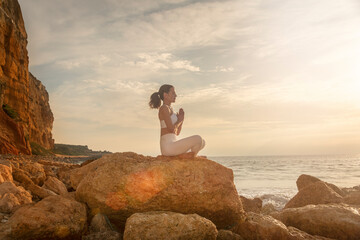 sporty woman practicing yoga and meditating on rocks by the sea at sunrise or sunset.