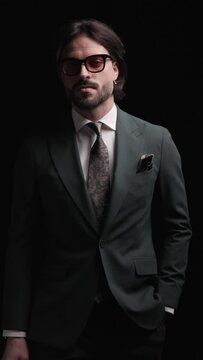 sexy unshaved businessman in elegant suit holding hands in pockets, walking in a confident way and smiling on black background