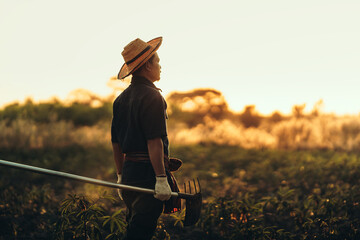 A cassava farmer holds a hoe and stands in the middle of his field during sunset.