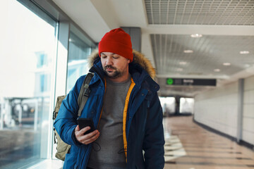 Young male traveler on an airport, wearing warm blue jacket and red knitted hat texting with his...