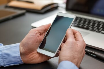 Closeup of hands holding and using a smartphone, with a laptop in the background on a desk at an office Generative AI