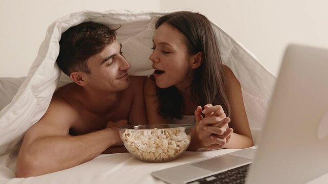 Intimate couple watching a movie on a laptop and sharing popcorn while lying in bed under a cozy duvet at night.