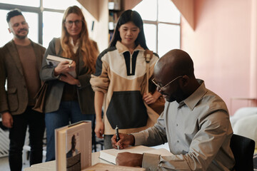 Young male writer signing items of his new published book bought by intercultural public during autograph session