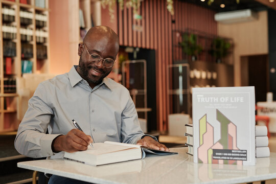 Happy young African American man in eyeglasses putting his signature on blank page of his new book while sitting by table