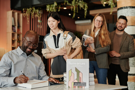 Young smiling writer signing first page of his book given him by one of readers standing in queue during autograph session