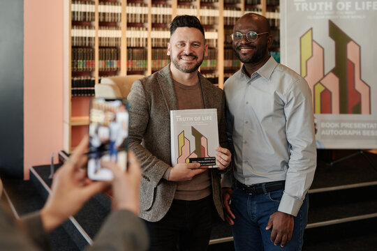 Two happy young intercultural men looking at smartphone camera while one of them holding new book of his favorite author