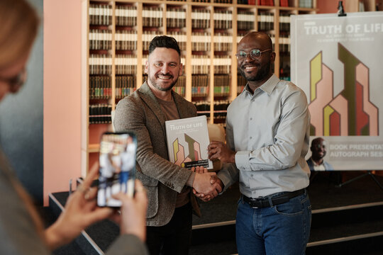 Young successful author shaking hand of his fan holding new book while both looking at smartphone camera during public event