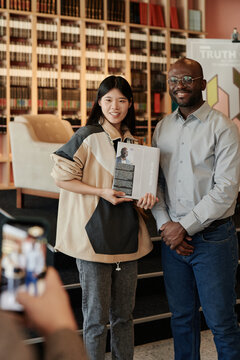 Happy young Asian woman with bestseller in hands standing by famous author while her friend taking picture of them on smartphone