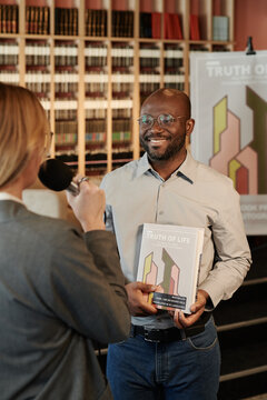 Happy young African American author with his new book in hands answering questions of journalist during presentation
