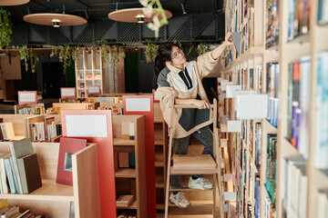 Young female assistant of spacious modern book shop searching for particular item on one of multiple shelves