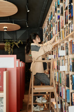 Side view of young female student or librarian standing on stepladder in front of huge stack with books and looking for particular one