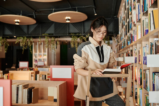 Young Asian female student in eyeglasses and casualwear looking through text in book while standing on stepladder in library
