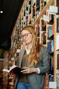 Young clever woman in eyeglasses holding open book and reading it while standing by huge stacks of shelves in library