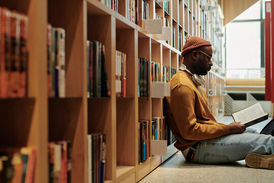 Young serious male student in casualwear reading book on the floor of college library while preparing for exam
