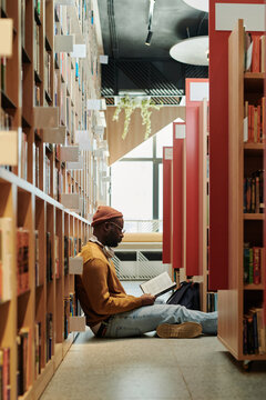 Side view of African American guy in casualwear sitting on the floor of library between high stacks of shelves with books