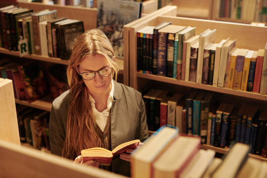 Girl with long blond hair looking through text in open book while standing between shelves with variety of novels in shop