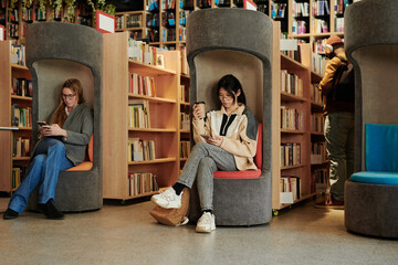 Two young intercultural women using smartphones in library or book shop against African American guy standing between shelves